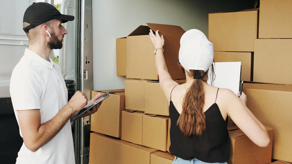A man and woman are inside the back of a moving van, surrounded by stacked brown cardboard boxes of various sizes, some open and partially empty. The man, wearing a white t-shirt, black cap, and wireless earbuds, is holding a tablet or clipboard while observing the packing process. The woman, dressed in a black sleeveless top and white cap, is reaching up to adjust or inspect the boxes. The interior of the van is well-lit, with natural light illuminating the scene, and a few packing materials such as plastic wrap or padding may be visible nearby. This image captures the home relocation process, including packing and loading furniture and boxes, as part of a professional removal service, with the focus on organized packing and careful handling typical of [COMPANY_NAME] and similar removal providers in Barking. The environment appears clean and tidy, emphasizing efficient furniture transport preparation in a residential setting.