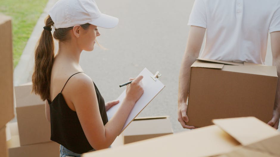 A young woman with long brown hair tied in a ponytail, wearing a black sleeveless top, a white baseball cap, and holding a clipboard with a pen, is standing outside a property on a paved driveway. She appears to be taking notes or conducting an inventory during a house relocation process. Behind her are several cardboard boxes, some sealed with packing tape, positioned on the ground and partially inside the open doorway of a house. To her right, a person wearing a white t-shirt is carrying a medium-sized cardboard box, which is held securely with both hands. The scene is well-lit with natural daylight, indicating daytime, and shows part of a vehicle or van used for furniture transport off to the side. This setting reflects a typical packing and loading process involved in professional removals by Man and Van Barking, supporting efficient and secure home relocation and furniture transport services as described in the webpage about avoiding hidden costs in Barking removals.