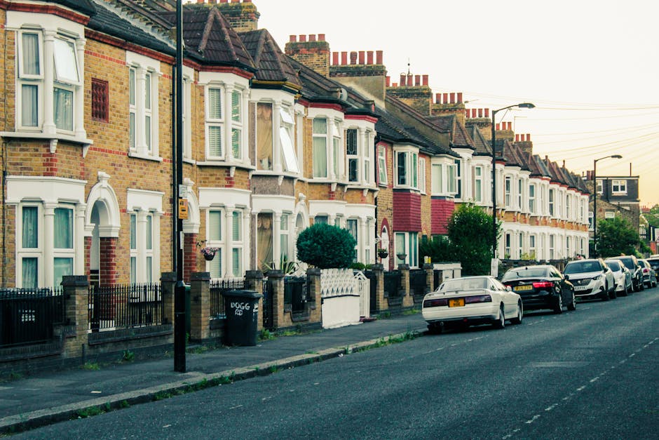 A row of terraced houses with brick facades and white bay windows, situated along a residential street in Barking, with a line of parked cars including a white coupe, a black sedan, and a white SUV along the curb. The pavement features a black lamppost and a black wheelie bin labeled 'DOGG', while a small green bush is visible in front of one house. The houses have front gardens enclosed by low brick and metal fences, some with potted plants and decorative elements. The street is quiet with a clear sky and warm lighting, typical of an evening or early morning setting. This scene illustrates residential property arrangements and parking arrangements relevant to vehicle access during house removals or relocations, as handled by professional removals services like Man and Van Barking.