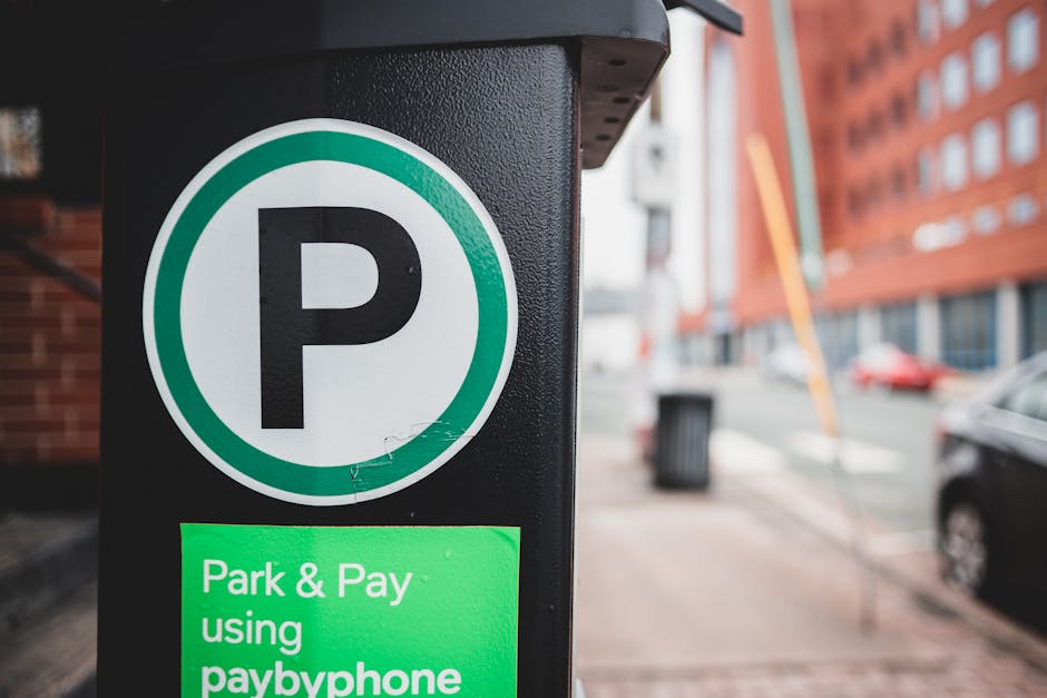 A close-up image of a black parking meter or payment terminal situated on a city pavement, displaying a green sign that reads 'Park & Pay using paybyphone' in white text. The photograph is taken during daytime with a blurred background showing a red brick building, parked cars, and street elements such as a trash bin and utility pole. The setting suggests an urban environment suitable for house removals or furniture transport, with the parking meter indicating adherence to local parking regulations. The scene highlights the importance of understanding parking rules in Barking and Dagenham for efficient home relocation or moving services, and the equipment and environment imply logistical planning involved in moving and packing processes. Man and Van Barking occasionally references such elements to support the context of house removals and moving logistics.