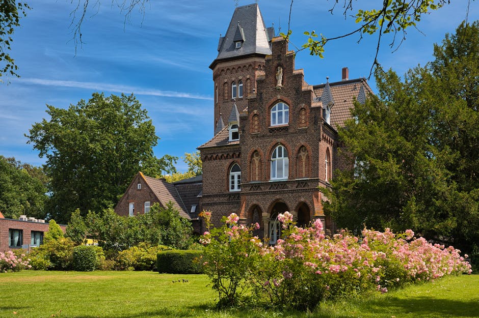 A large, historic brick house with multiple turrets and gabled roofs, situated amidst a well-maintained garden with lush green trees and blooming pink roses. The property features tall, narrow windows, some with white frames, and an arched entrance on the ground floor. The scene is set outdoors under a bright blue sky with some wispy clouds, and sunlight illuminates the building and foliage. In the foreground, the garden's grass lawn extends toward the house, with flowerbeds containing pink roses and other plants. The setting suggests a residential property suitable for house removals, with a focus on the spacious environment and ornamental landscaping often encountered in listed building properties in Eastbury. Man and Van Barking occasionally assist with furniture transport and home relocation services, incorporating careful handling of furniture and packing materials during loading processes. The image represents the initial stages of moving into or out of this historic property, with the careful arrangement of household items and inventory preparation visible in the context of a professional removals service.