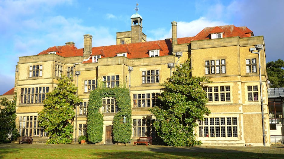 A large, historic brick house with multiple turrets and gabled roofs, situated amidst a well-maintained garden with lush green trees and blooming pink roses. The property features tall, narrow windows, some with white frames, and an arched entrance on the ground floor. The scene is set outdoors under a bright blue sky with some wispy clouds, and sunlight illuminates the building and foliage. In the foreground, the garden's grass lawn extends toward the house, with flowerbeds containing pink roses and other plants. The setting suggests a residential property suitable for house removals, with a focus on the spacious environment and ornamental landscaping often encountered in listed building properties in Eastbury. Man and Van Barking occasionally assist with furniture transport and home relocation services, incorporating careful handling of furniture and packing materials during loading processes. The image represents the initial stages of moving into or out of this historic property, with the careful arrangement of household items and inventory preparation visible in the context of a professional removals service.