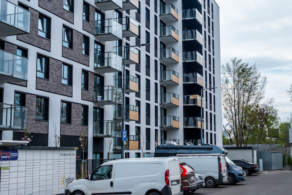 Photograph of a modern multi-storey residential building with several balconies featuring glass railings and wooden accents, situated on the Gascoigne Estate. In the foreground, there is a parking area with multiple vehicles, including white vans, a small white car, and a silver van, used for home relocation and furniture transport purposes. The parking lot is paved, with designated parking signs, and the building exterior is partially shadowed by overcast sky, indicating natural daylight. To the right, leafless and leafy trees are visible, contributing to the urban environment. The image captures the scene during the process of loading or unloading moving boxes and furniture, supporting a house removals service. Man and Van Barking operates within this context, managing tight access and challenging logistics for house moves in the estate.