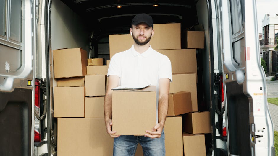A man wearing a white polo shirt, jeans, and a black cap is standing inside the open back of a white moving van, which is parked outside a residential property. He is holding a cardboard box with both hands and facing the camera. The van's interior is filled with neatly stacked cardboard boxes of various sizes, some wrapped in plastic and others simply stacked on top of each other, suggesting the process of packing and furniture transport during a home relocation. The van is positioned on a paved driveway beside a house and garden area, with natural daylight illuminating the scene. Nearby, additional boxes and packing materials are visible, indicating an ongoing moving process handled by professionals such as Man and Van Barking, specializing in removals and tight access moves like those on the Gascoigne Estate. The image captures the loading process, highlighting careful handling of boxes and the logistics involved in house removals.
