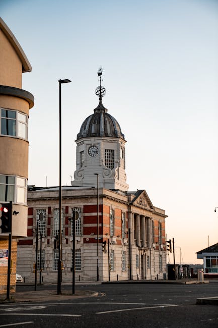 A historic white and red brick building featuring classical architectural details, with a domed clock tower topped by a weather vane, visible during daytime with clear skies. In the foreground, there are a few traffic lights, street lamps, and a paved road, indicating an urban setting in Barking. The surrounding area includes modern and older buildings, with parts of a residential property seen on the left side. The scene captures the exterior environment where house removals and furniture transport services by Man and Van Barking may operate, highlighting the importance of careful logistical planning for moving within the town centre. This image exemplifies the typical architecture and streetscape that professionals involved in house relocations might navigate during a comprehensive packing and moving process, ensuring safe transportation of personal belongings and furniture to new addresses.