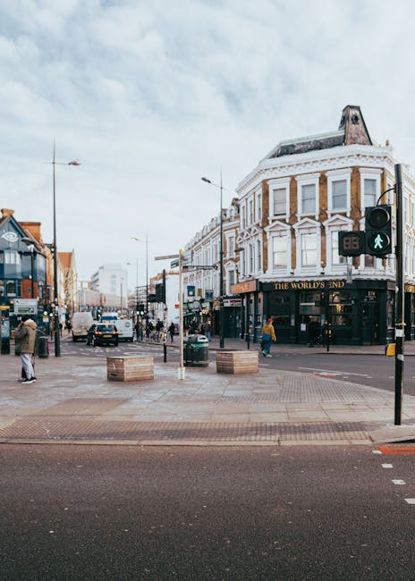 A street scene in Barking town centre showing pedestrian crossing with a green pedestrian light active. To the right, there is a three-story corner building with white decorative architectural elements and a sign reading 'The World's End' on the ground floor, likely a pub or local business. Several pedestrians are walking across the intersection, some with shopping bags, while others wait at the curb. On the left side, there are additional commercial buildings with shop fronts, and vehicles, including cars and vans, are visible in the background. The street is lined with lampposts and street furniture, including large wooden planters. The sky is partly cloudy, and the overall scene suggests a busy urban environment suitable for residential relocations, with potential activity involving moving furniture and boxes, consistent with house removal services provided by companies like Man and Van Barking.