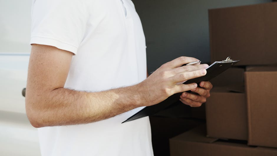 Close-up of a person wearing a white t-shirt holding a black clipboard and writing with a silver pen, standing in front of a stack of cardboard boxes and furniture in an indoor setting. The boxes are sealed with brown packing tape, and some are partially covered with protective plastic wrap. Several cardboard boxes are placed on the floor, indicating a packing or unpacking process during a home relocation. The scene shows the individual engaged in organizing or documenting items before transportation, with natural lighting highlighting the workspace. The environment appears to be inside a house or apartment, and the background includes a door and neutral-toned wall. This image reflects the planning and execution phase of a professional removals service, such as those offered by Man and Van Barking, related to packing and moving logistics involved in house removals.