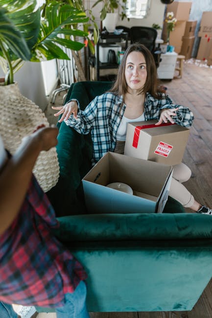 A young woman with long brown hair, wearing a blue and white checked shirt, is seated on a green sofa inside a home during a packing process. She appears to be engaged in a conversation with another p