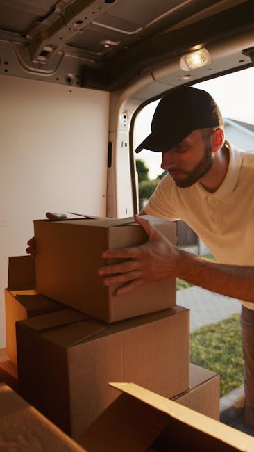 A man wearing a black cap and a white T-shirt is inside the back of a van, surrounded by stacked cardboard moving boxes of various sizes, some with open flaps. He is in the process of loading or unloa