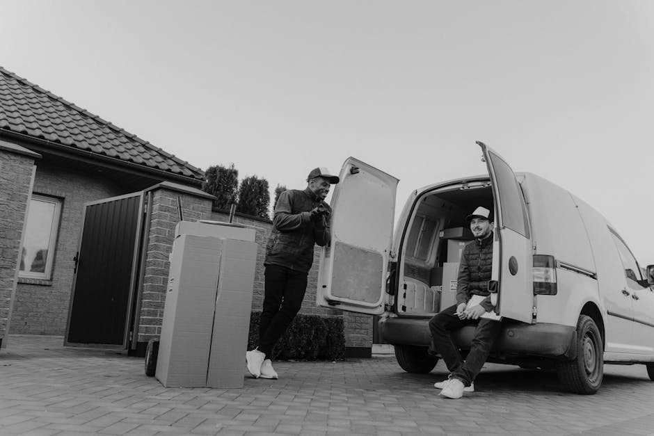 Two men are engaged in a home relocation process outside a residential property, with one standing on the driveway and the other sitting on the open rear doors of a white cargo van belonging to Man and Van Barking. The man standing is wearing a dark jacket, trousers, and a cap, and is holding a mobile phone, appearing to communicate or check details. The seated man is dressed in a quilted jacket, trousers, and a hat, smiling while resting his hands on his knees. The rear of the van is open, revealing a partially visible interior with packed boxes, some wrapped in plastic or cardboard, and a small piece of furniture or appliance partially loaded inside. Nearby, on the paved driveway, there are more moving boxes and a large, upright cardboard box ready for transport. The surrounding environment includes a brick house with a tiled roof, a small garden area, and a black fence, with natural daylight illuminating the scene. This image illustrates furniture transport and packing during a house move, reflecting the logistics involved in a same-day moving service, typical of [COMPANY_NAME]'s removals work as seen on the [PAGE_TITLE] page.