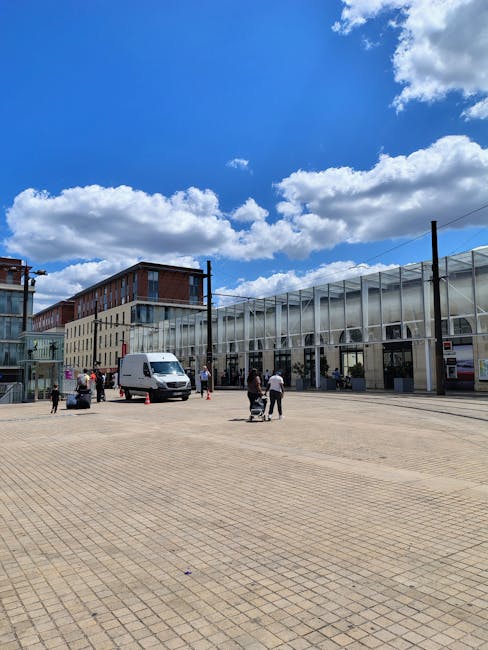 A wide outdoor view of a modern urban building with a glass facade and steel framework, situated behind a large, paved open area under a bright blue sky with scattered white clouds. Several people are visible in the scene, some walking and others standing near a white delivery van parked close to the building entrance. The scene depicts a typical day of home or commercial relocation activity, with individuals preparing or coordinating the transportation of furniture and boxes, supported by the services of Man and Van Barking for efficient removals and moving logistics. The environment features a combination of contemporary architecture and open space, suitable for loading and unloading items during packing and moving processes.