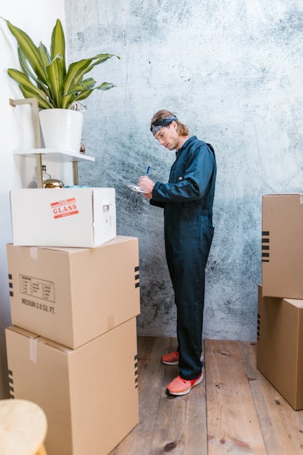 A woman with curly brown hair, wearing a grey cardigan and white top, is seated indoors taking notes on a notepad with a black pen, amidst packing materials such as an open cardboard box and a green-t