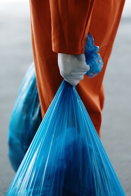 A person dressed in orange work overalls and white shoes is standing inside a property, holding two large blue plastic trash bags filled with household waste, presumably during a home relocation or pa